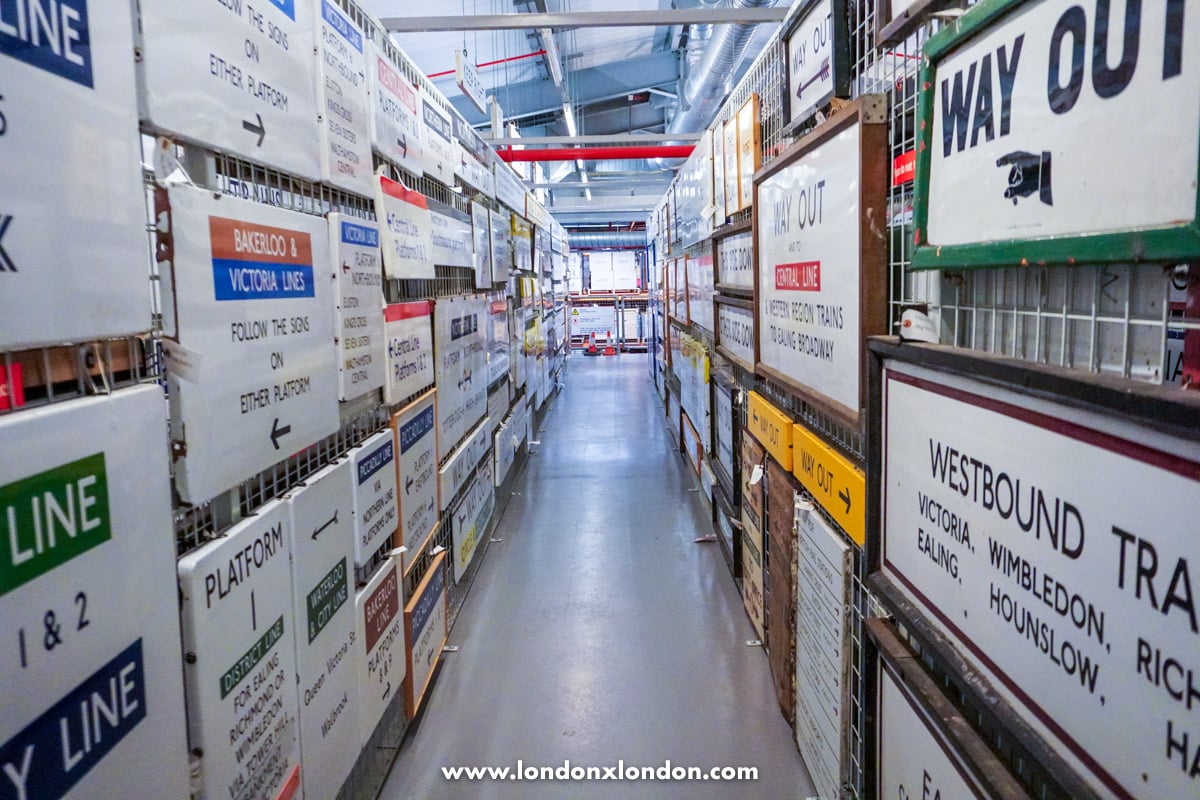 A narrow aisle between storage racks packed with old Underground station signs and platform boards