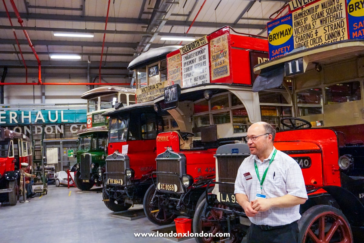 A guide standing beside a row of vintage red London buses inside the Museum Depot's hall