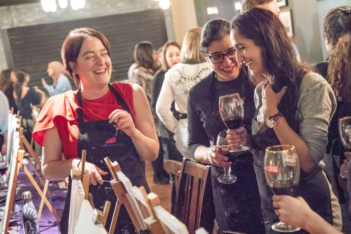 Three women laughing together holding wine glasses at a paint and draw class, easels and other painters behind them