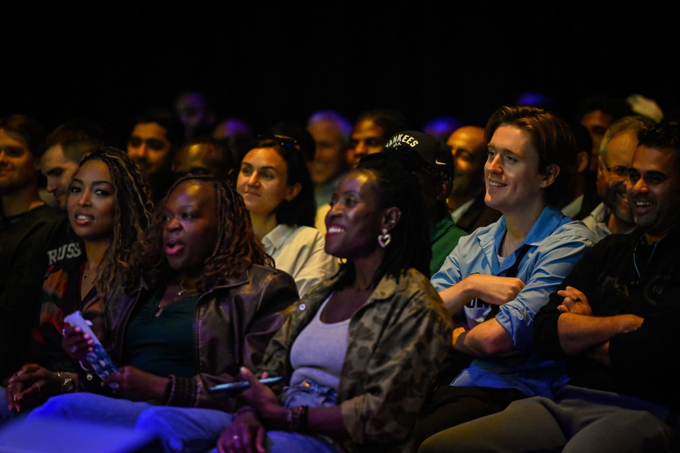 A packed audience in a darkened auditorium at Kings Place, faces lit by the stage, laughing and reacting