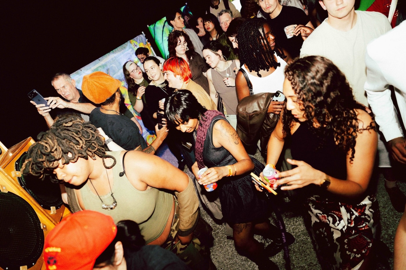 A packed crowd dancing beside a wooden sound system speaker at a V&A East late night, drinks in hand and a phone held up to film