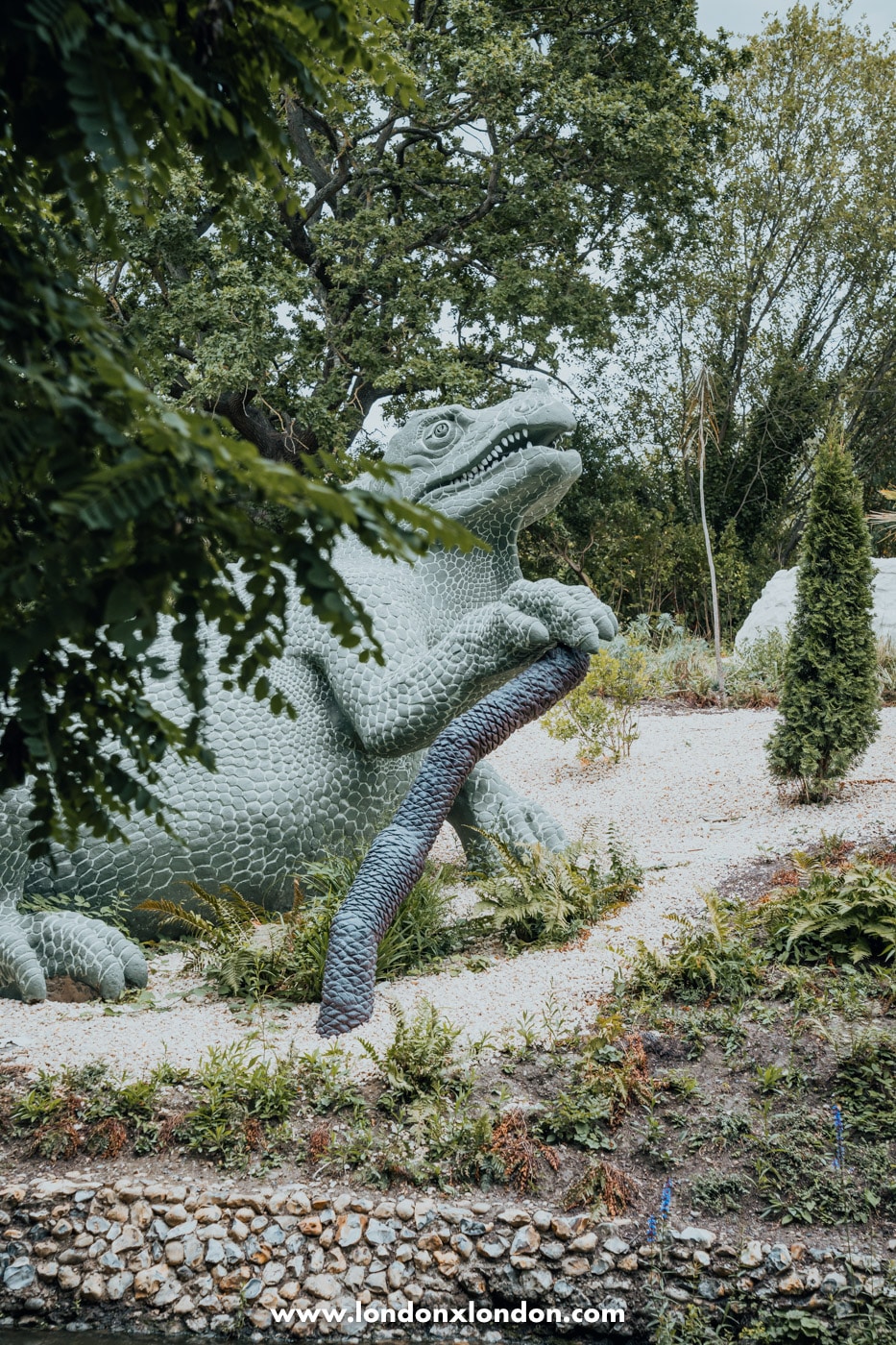 A restored Iguanodon sculpture at Crystal Palace Park, repainted in green with its tail curving down the bank