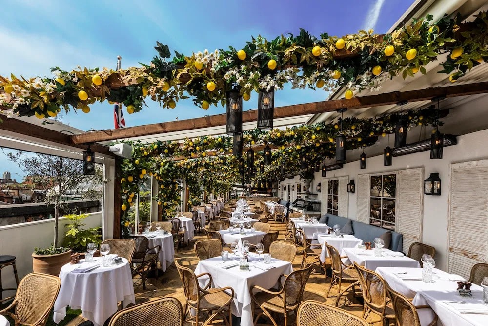 Rattan chairs and white-clothed tables under alto by San Carlo's summer installation, with blue sky above the Selfridges rooftop