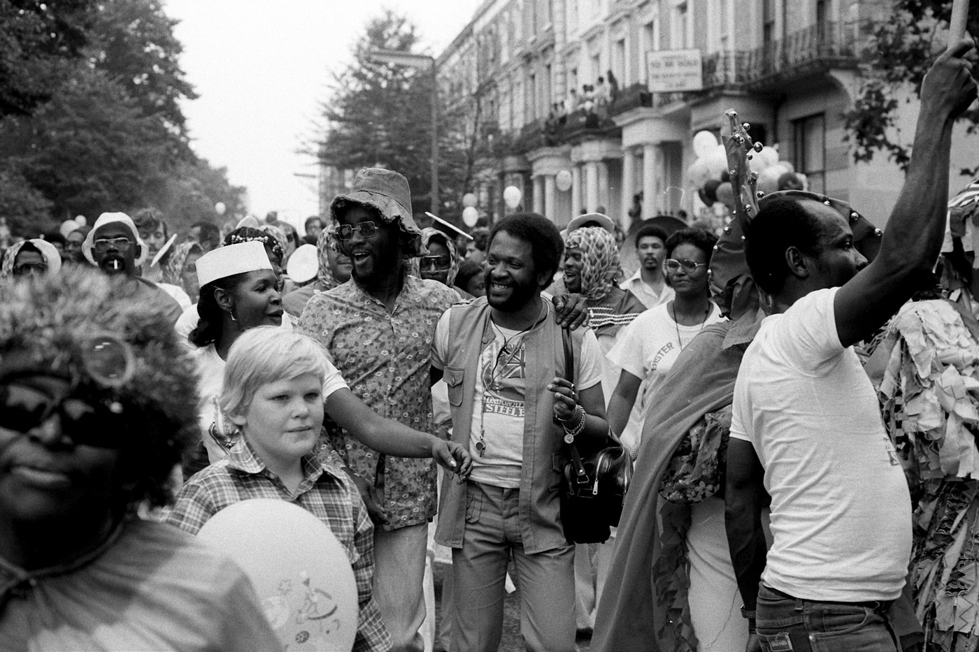 A black-and-white photograph from Notting Hill Carnival in 1976, showing a crowd walking down a residential street