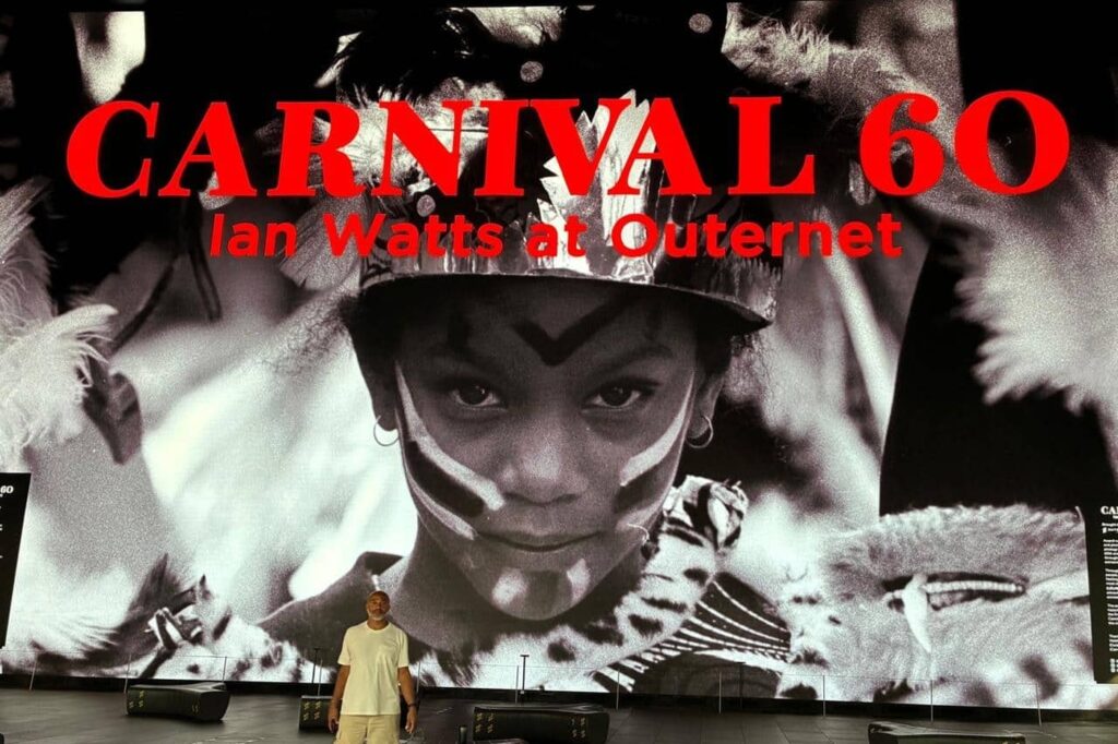 The 'Carnival 60: Ian Watts at Outernet' title screen, showing an archive black-and-white photograph of a young Carnival-goer in feathered costume