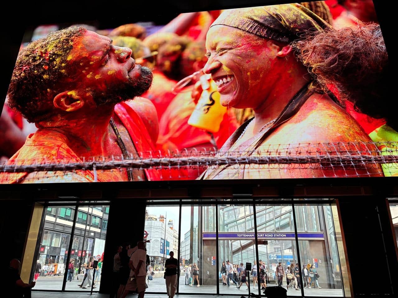 Two Carnival-goers covered in coloured powder pictured on the giant screens at Outernet London, with Tottenham Court Road station visible below
