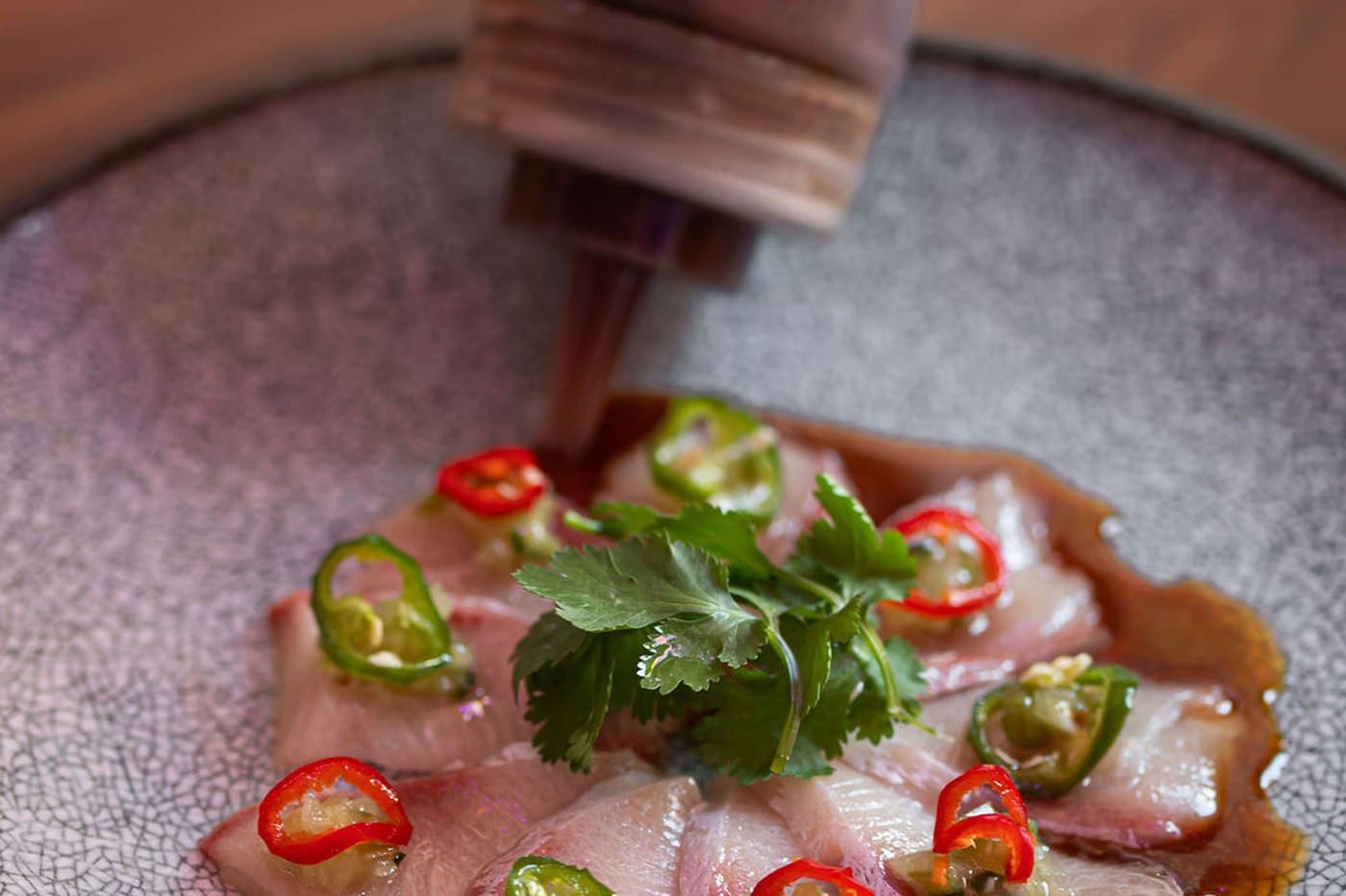 Slices of yellowtail hamachi sashimi on a grey plate with red and green chilli and coriander, ponzu poured tableside at Freak Scene in Balham.