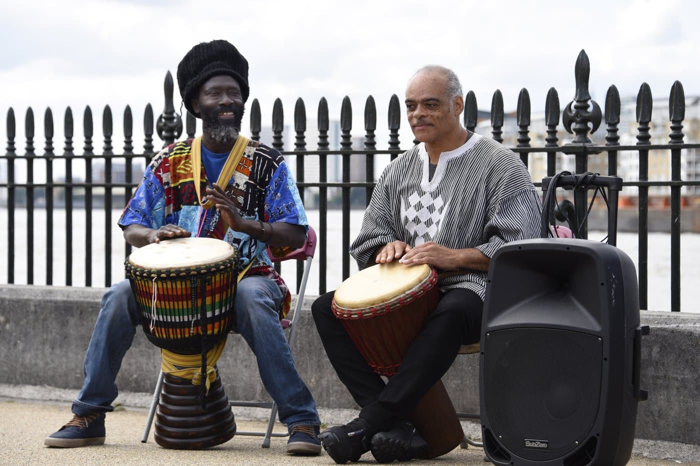 Two men play djembe drums beside railings overlooking the River Thames in Greenwich