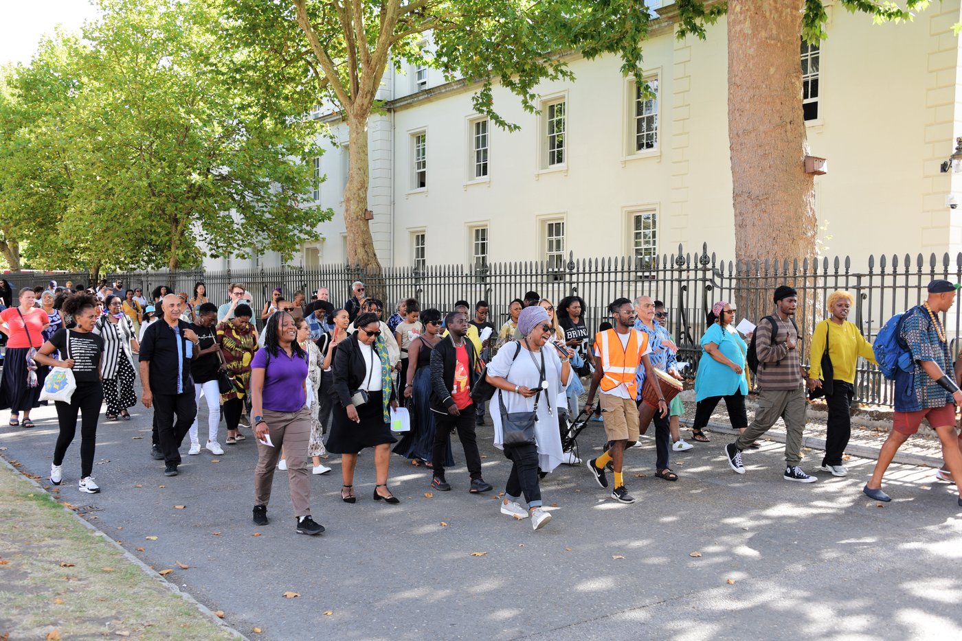 A crowd of people, including a drummer and a steward in a hi-vis vest, walk together along a path beside the Queen's House in Greenwich