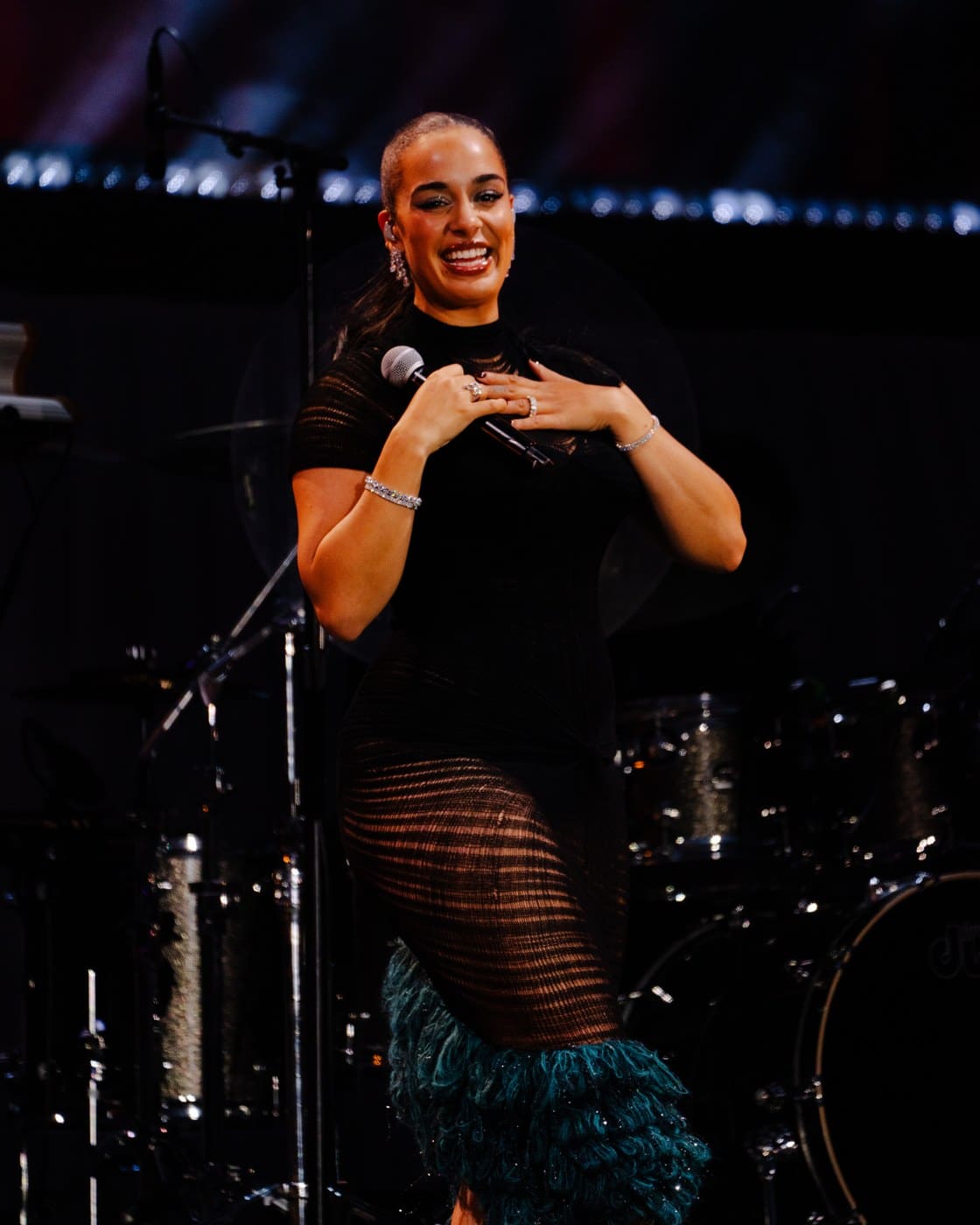 Jorja Smith smiling with both hands held to her chest on stage, in a black knit dress with teal feather trim
