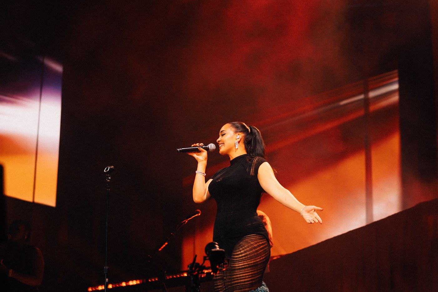 Jorja Smith singing into a handheld mic on stage, arm outstretched, against a deep orange backdrop