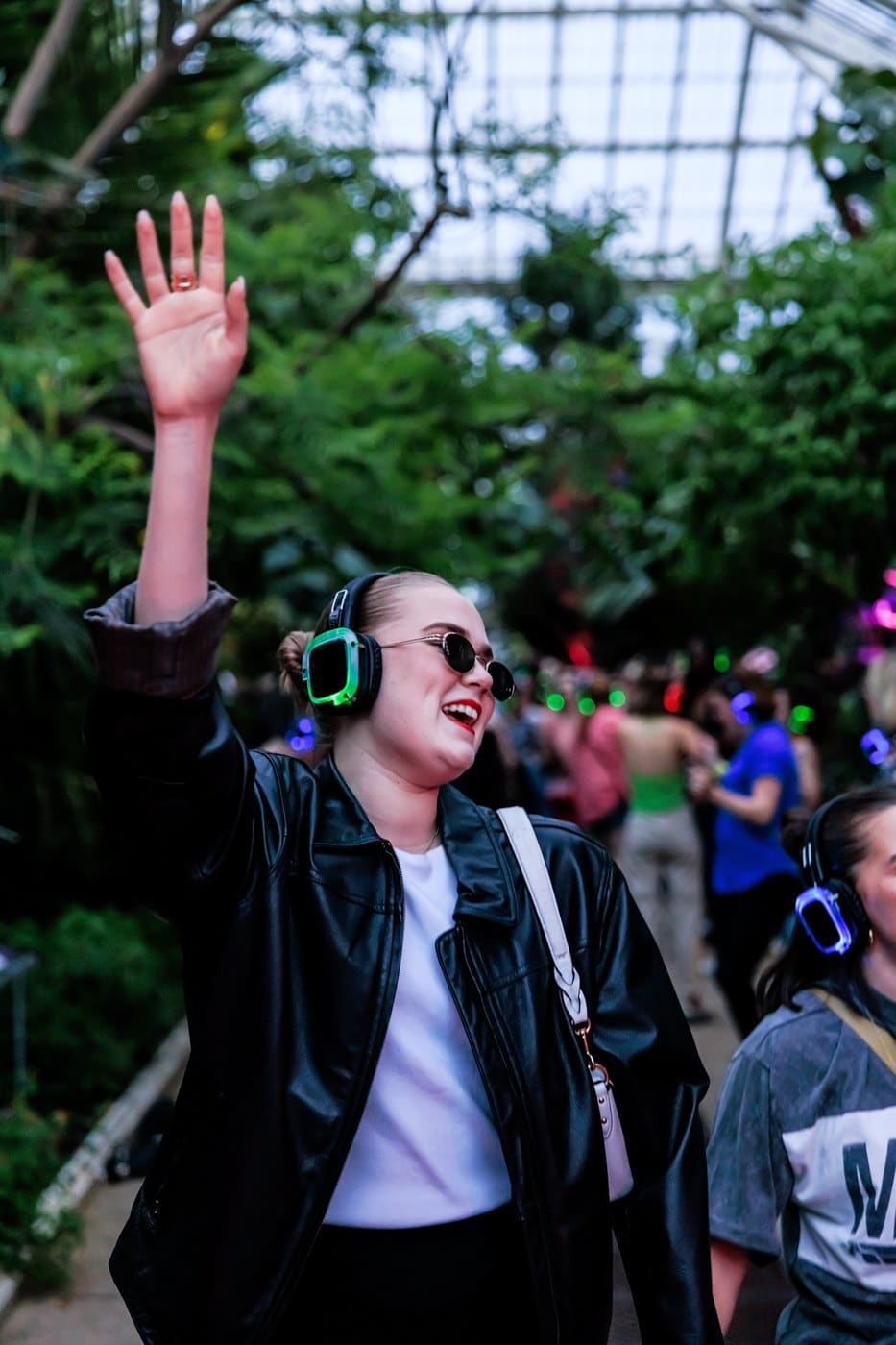 A woman in sunglasses and green-lit headphones dancing with one arm raised inside the Temperate House