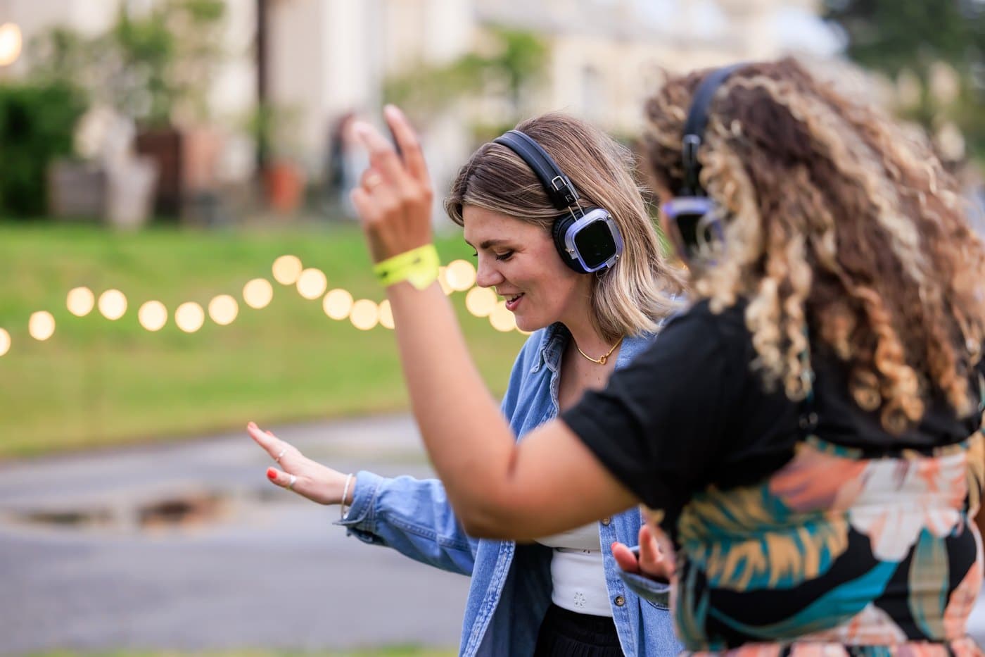 Two women in headphones dancing on the lawn at Kew beneath a string of festoon lights