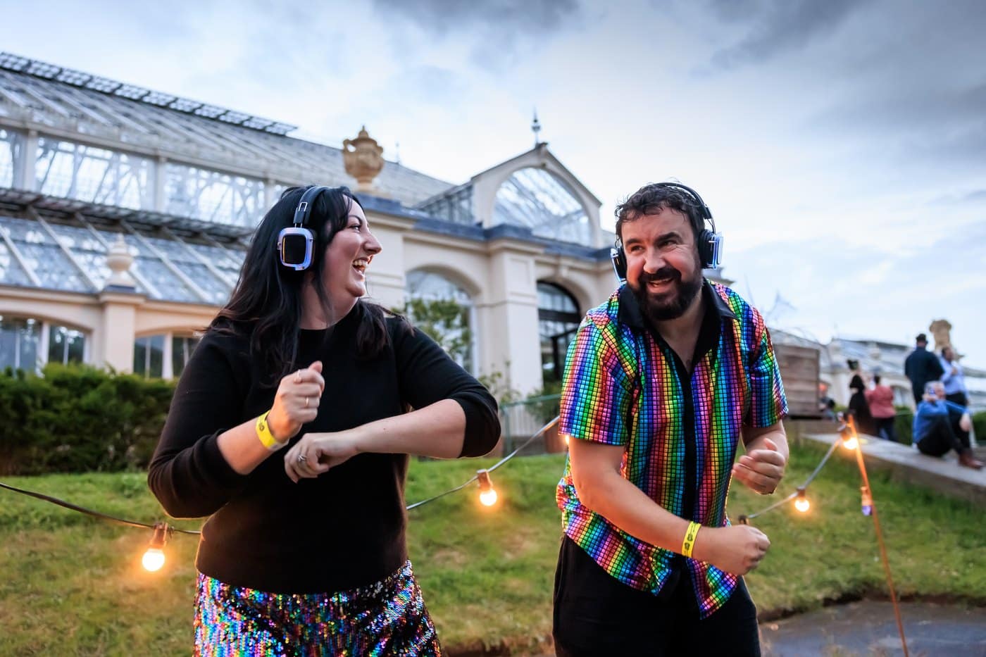 Two people in headphones laughing and dancing outside the lit facade of Kew's Temperate House