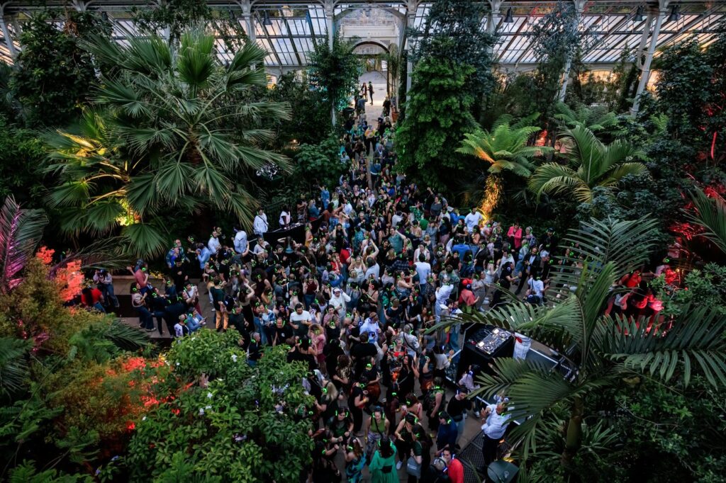 Hundreds of people wearing glowing headphones dancing between palms and tree ferns inside Kew's Temperate House at dusk