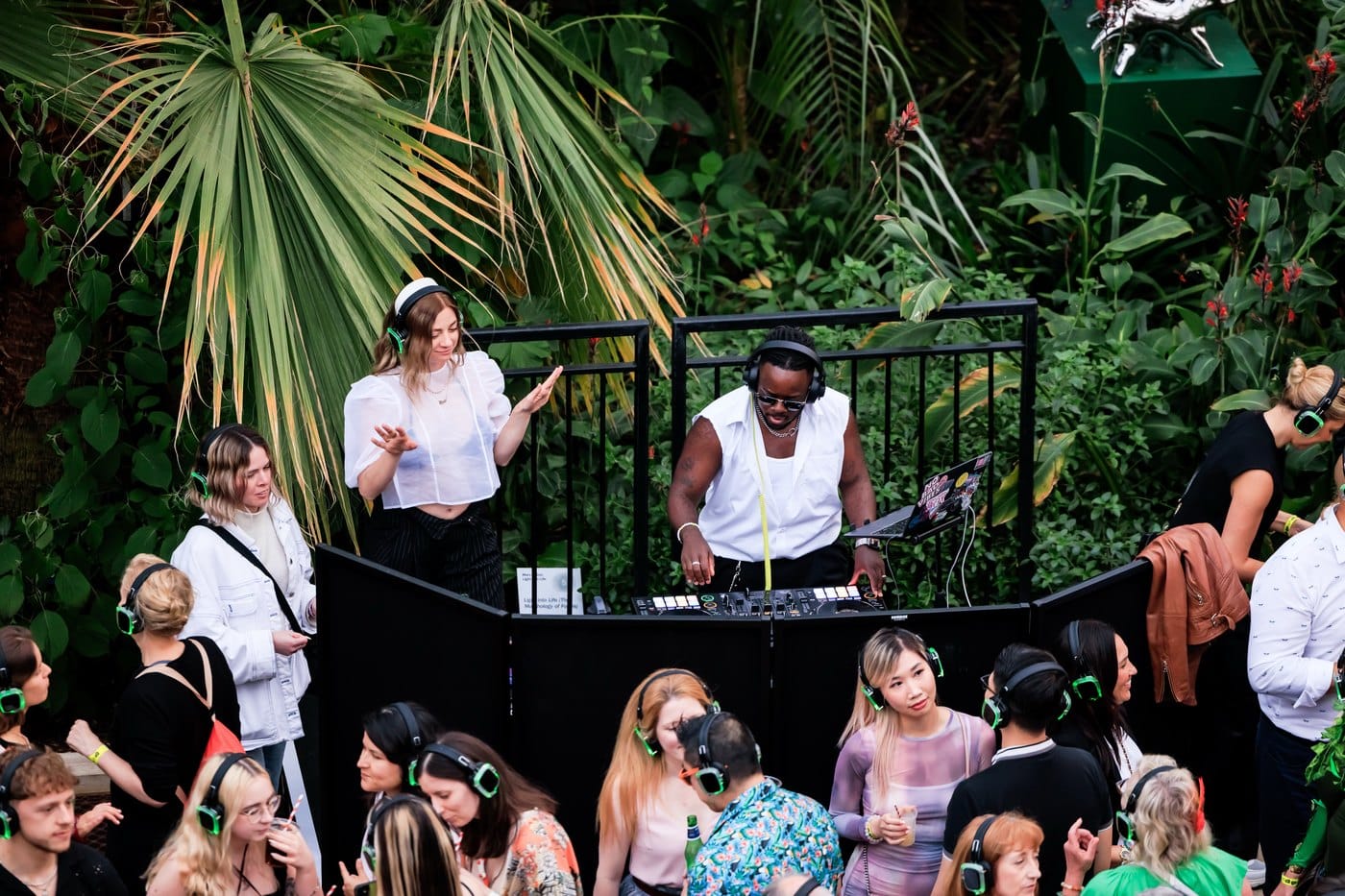 A DJ playing to a crowd in glowing green headphones beneath palm fronds inside Kew's Temperate House