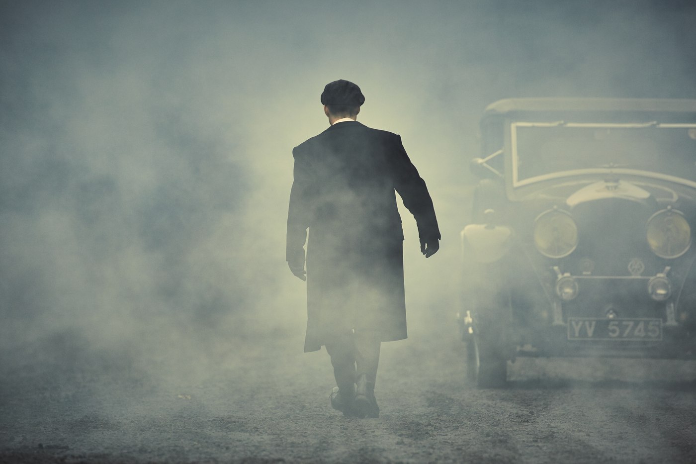 A Peaky Blinders figure in a long overcoat and flat cap walking away through smoke past a vintage motor car.