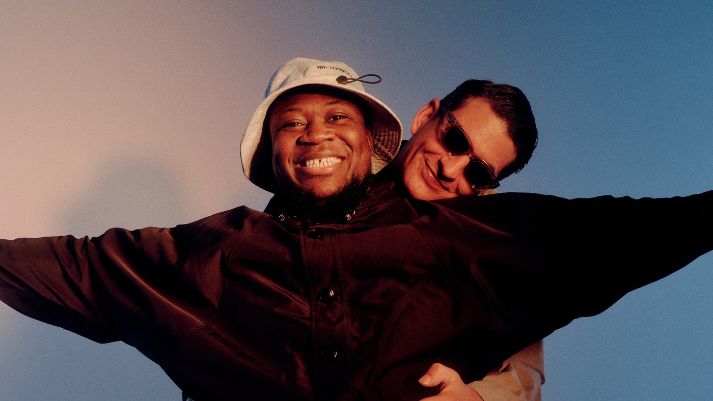Skream and Benga photographed arm in arm against a dusk sky, one in a white bucket hat and the other in sunglasses