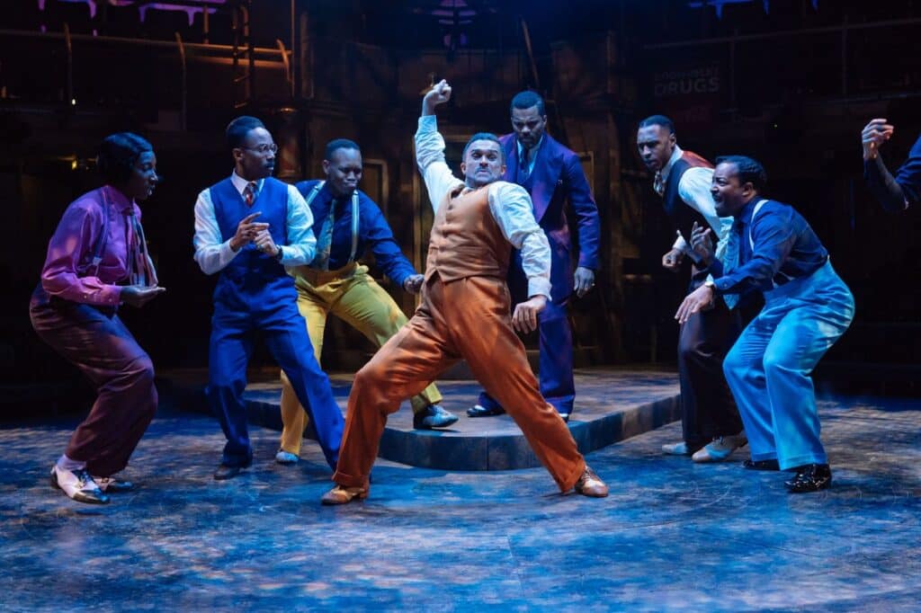 Eight performers in 1940s suits and braces mid-dance on a Talawa stage under blue light