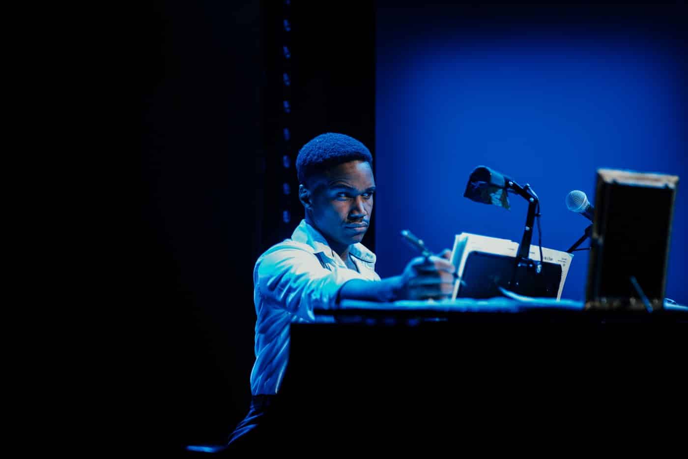 An actor at an upright piano in deep blue stage light, pen in hand, looking out to the audience