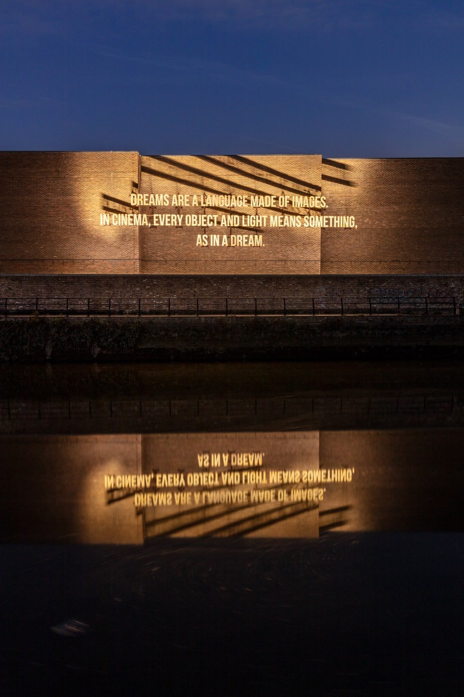 Zineb Sedira's illuminated text artwork Dreams Are a Language Made of Images, projected on a canal-side wall at 3 Mills Studios, reflected in the water