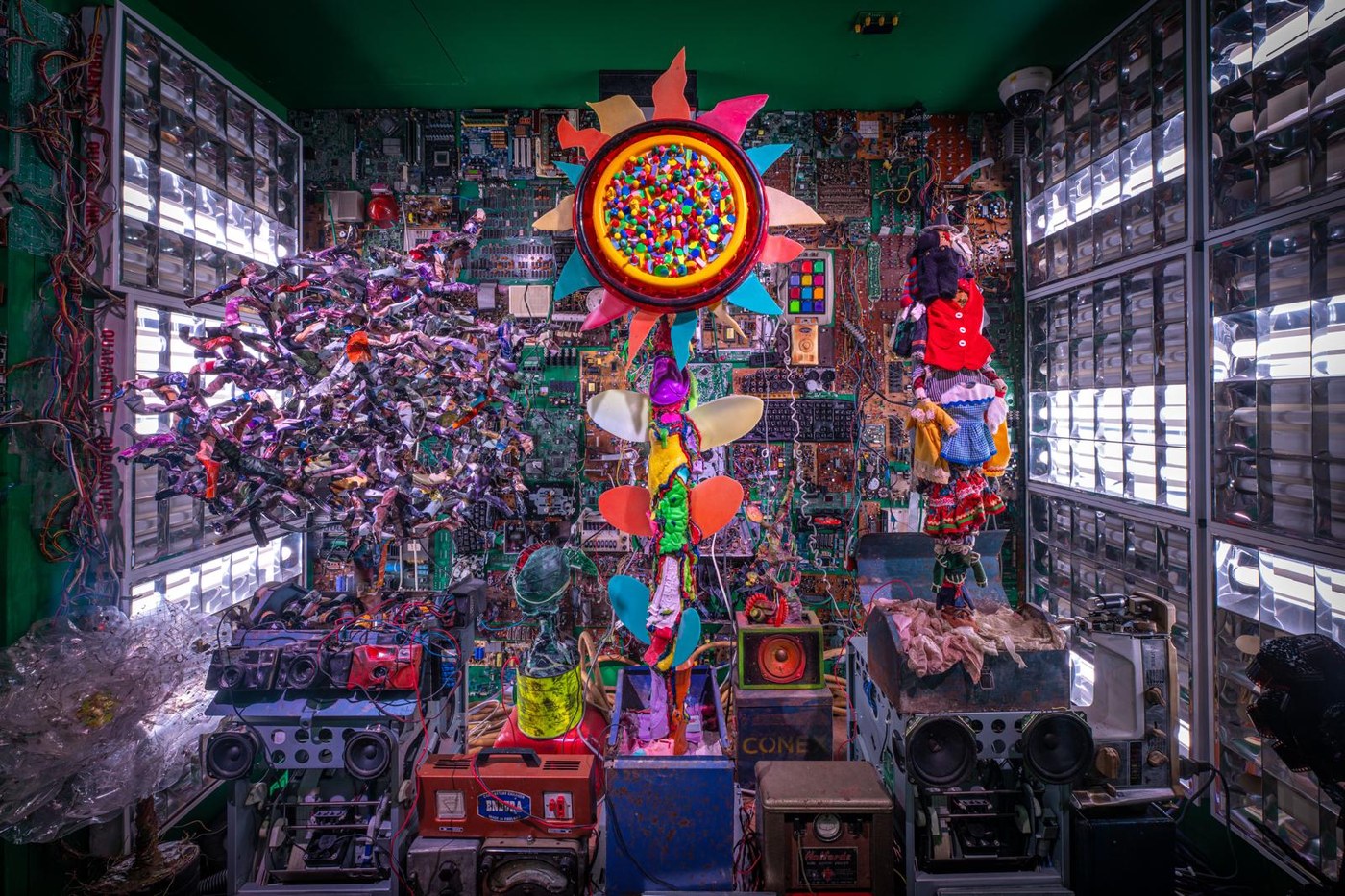 A room lined with circuit boards and light panels, with a large flower made from bottle tops and offcuts at its centre