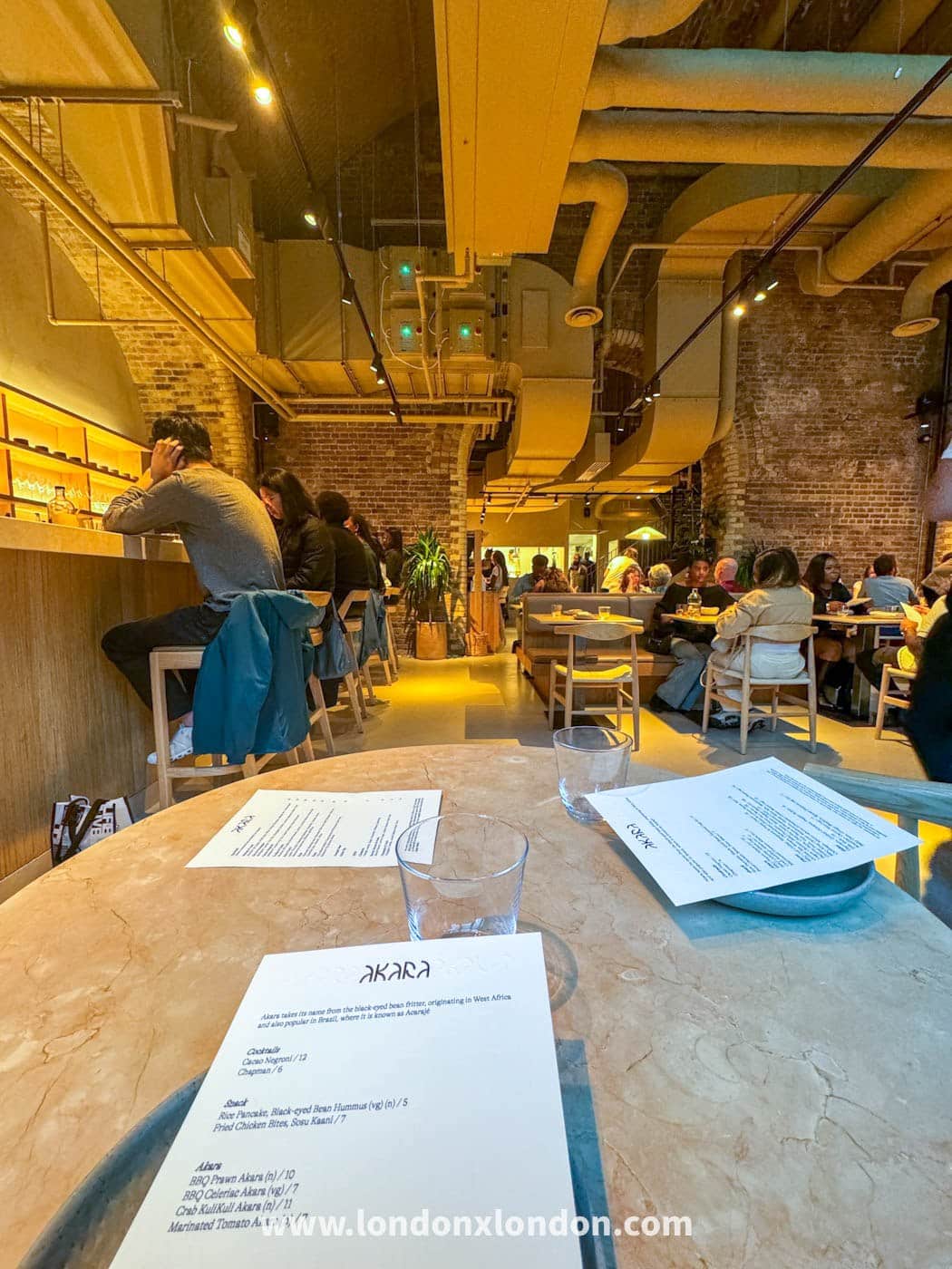 The dining room at Akara in Borough Yards, a brick railway arch with exposed ducting, counter seats along one side and tables opposite, with an Akara menu card on a marble table in the foreground