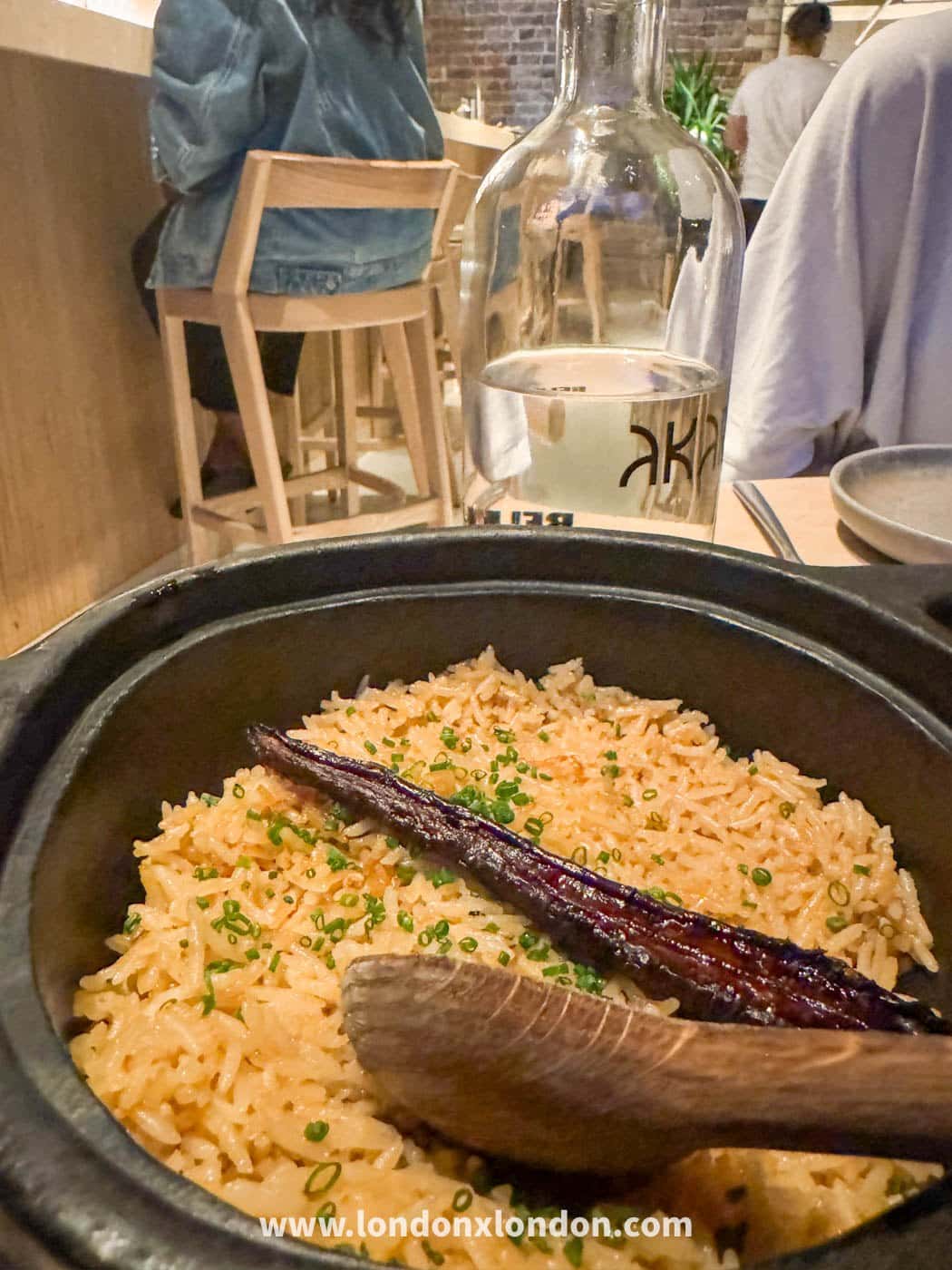 A cast-iron pot of orange-tinted rice scattered with chopped spring onion, a long charred aubergine laid across the top and a wooden spoon resting in it, with an Akara-branded water carafe behind