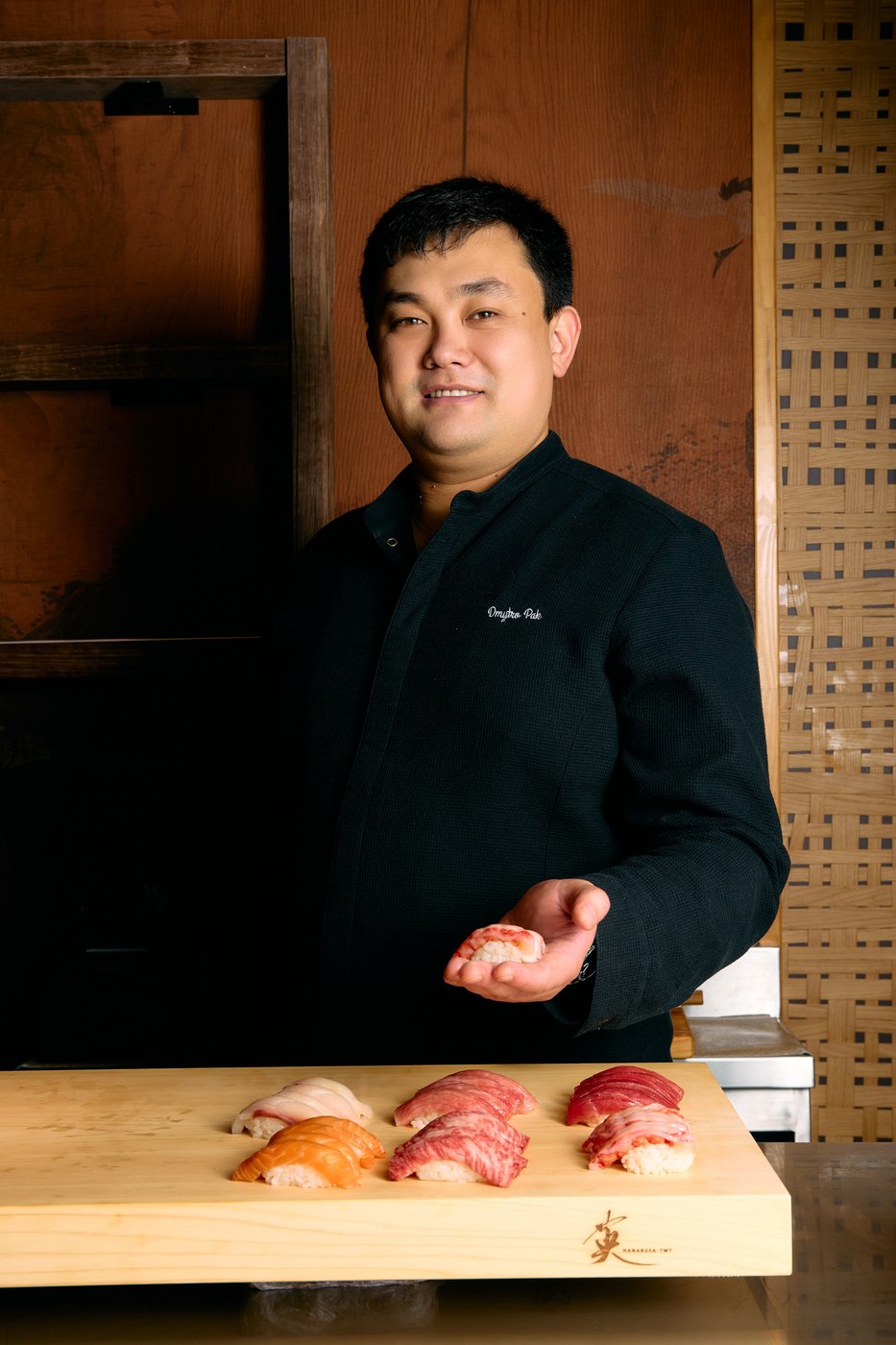 Creative chef Dmytro Pak in black chefs whites holding a single piece of nigiri in his palm behind a cypress counter set with six pieces