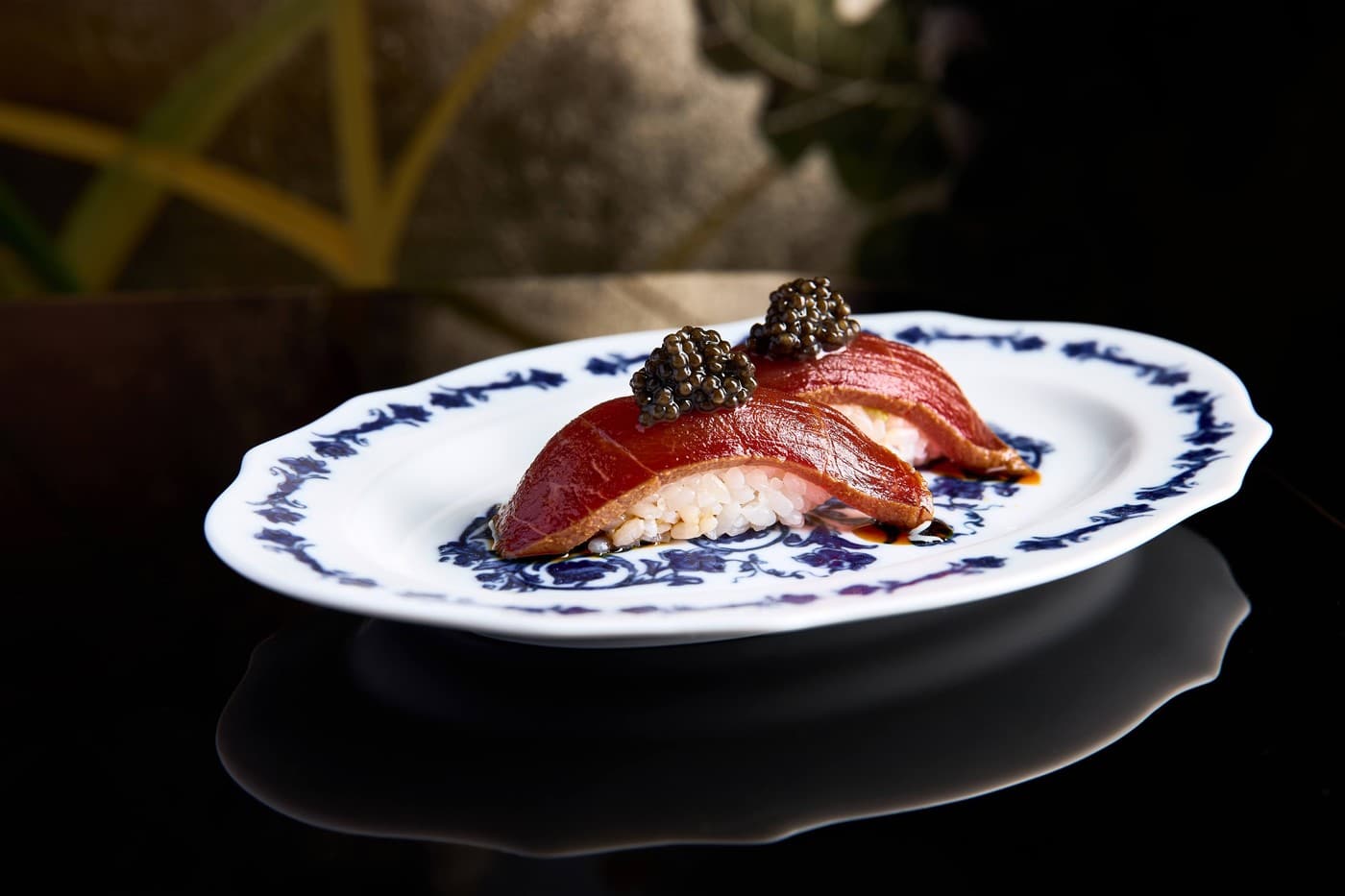 Two pieces of cured tuna nigiri each topped with a quenelle of black caviar, on a blue and white patterned plate