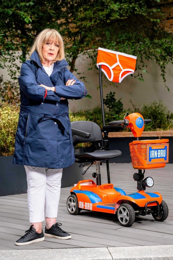 Amanda Barrie standing with her arms crossed beside an orange IRN-BRU mobility scooter with blue flame stripes and a pants-shaped flag