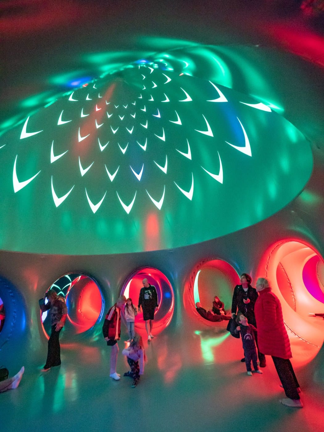 Visitors standing beneath a green glowing dome patterned with white chevrons inside ARBORIALIS