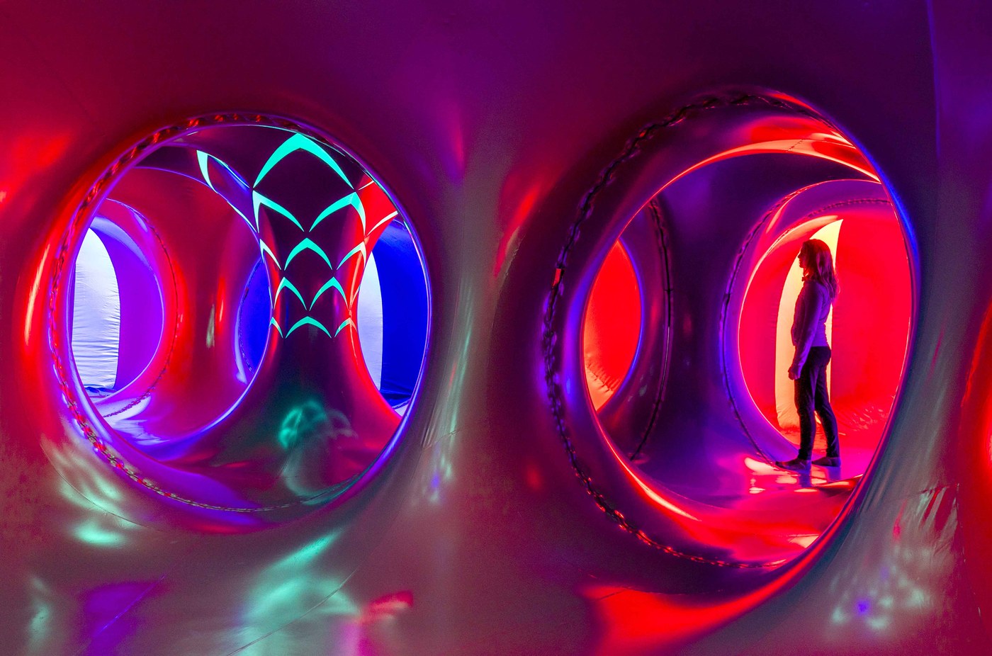 A visitor standing in a red glowing tunnel inside the ARBORIALIS Luminarium, a turquoise motif on the wall beside them