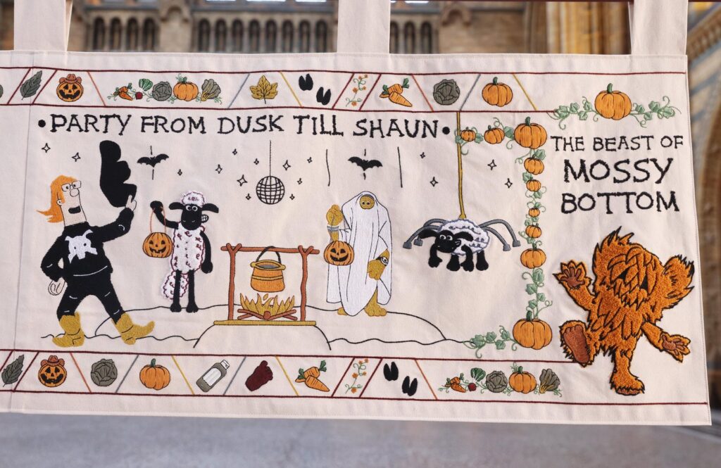 A panel of the Baa-Ewe Tapestry at the Natural History Museum showing Shaun the Sheep trick-or-treating at a farmyard Halloween party under the stitched caption Party From Dusk Till Shaun
