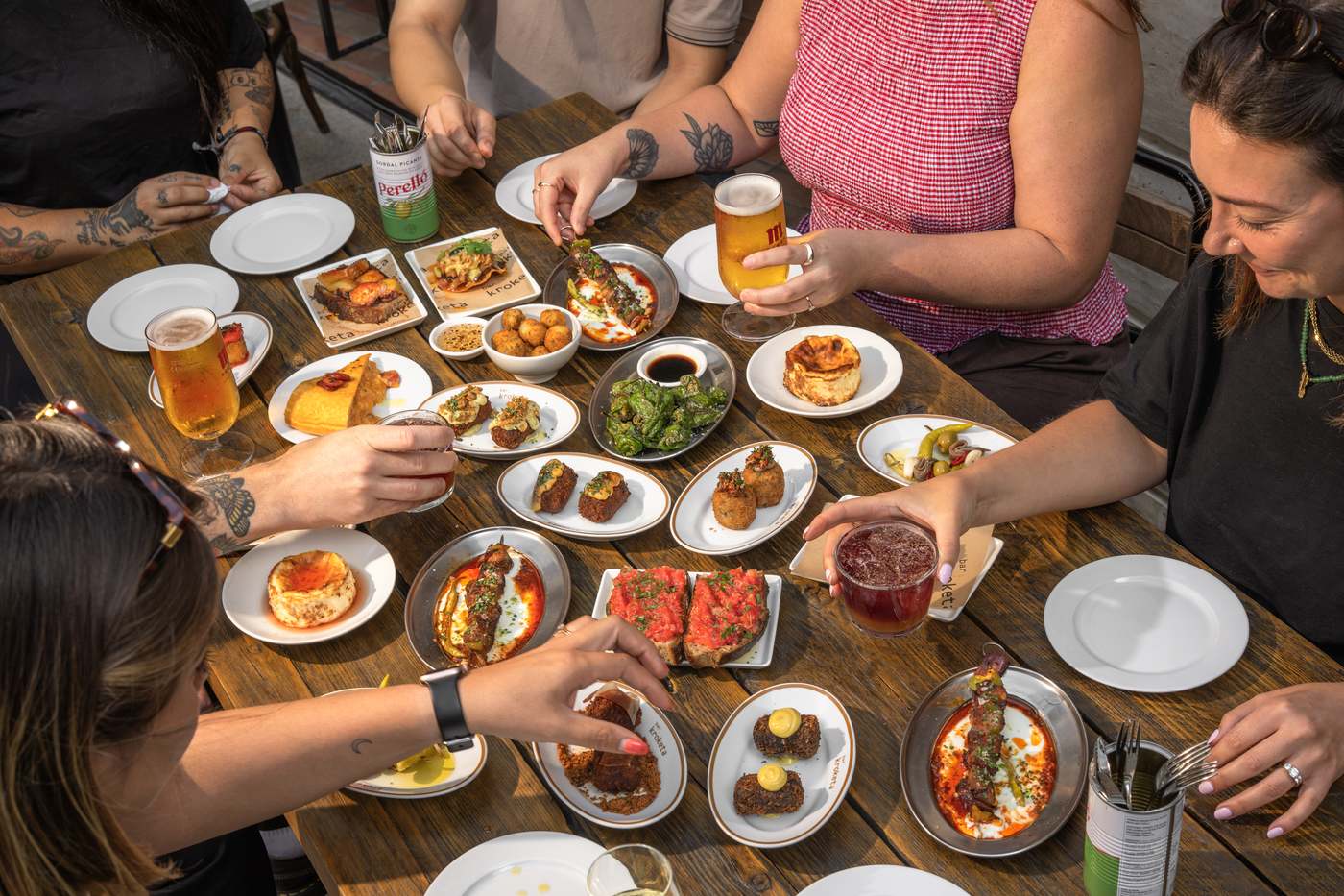 A packed table of croquetas, gildas, padron peppers and Basque cheesecake in late afternoon light, with hands reaching from every side