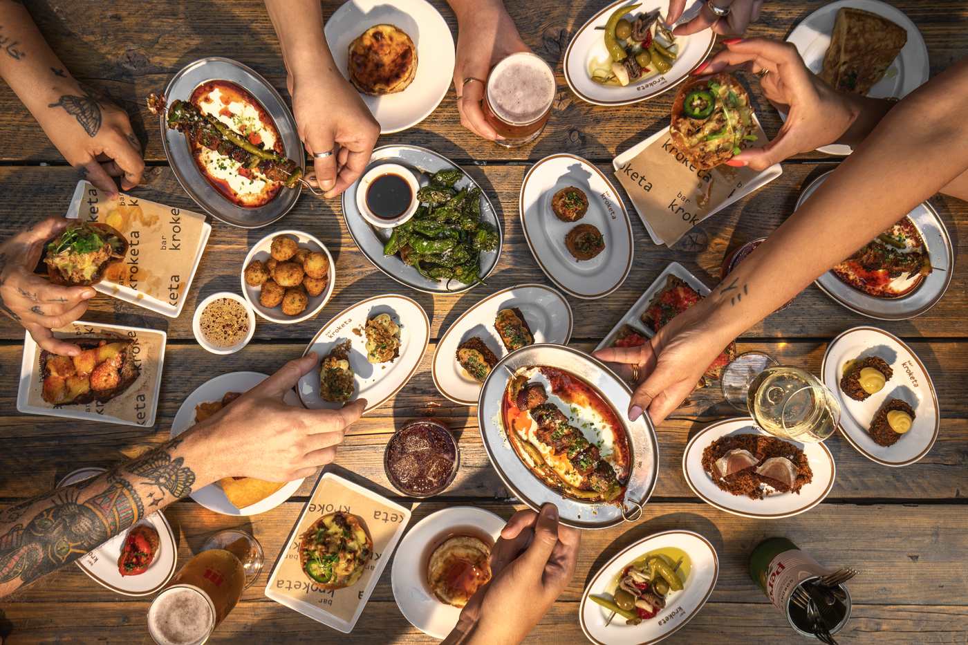 An overhead view of a wooden table crowded with croquetas, padron peppers, pan con tomate, Basque cheesecake and glasses of beer, with several people reaching in