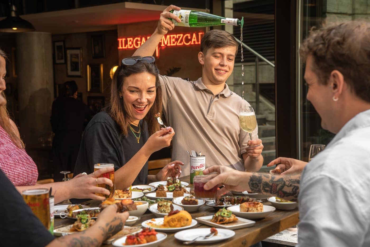 A man pouring txakoli from a green bottle held high above the table into a glass, while a woman laughs beside him