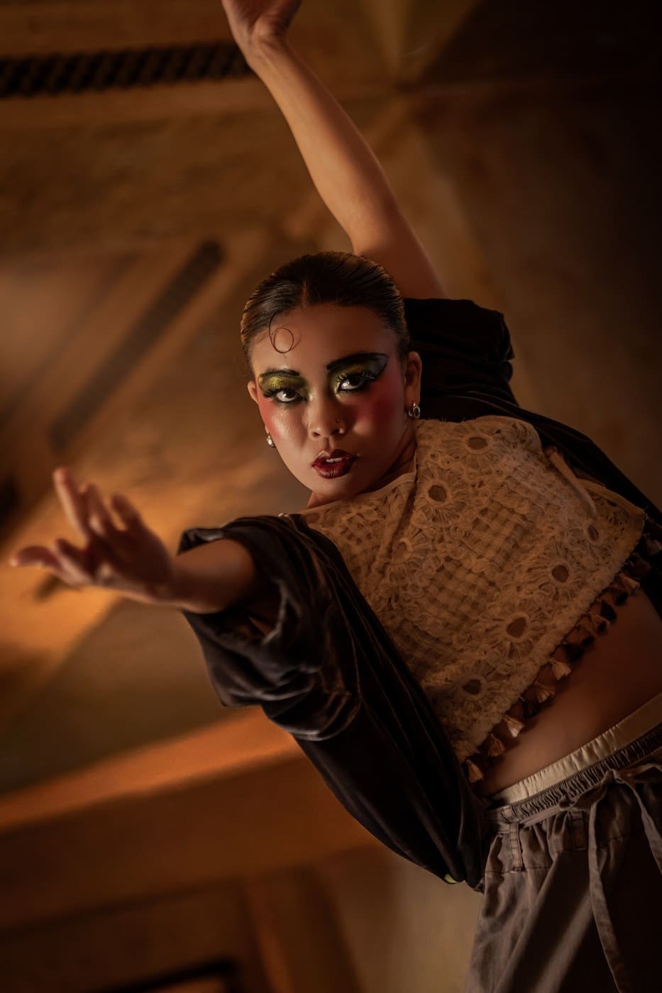 A Cabaret dancer in gold and green Kit Kat Club make-up, one arm raised, under the timber beams at Balthazar