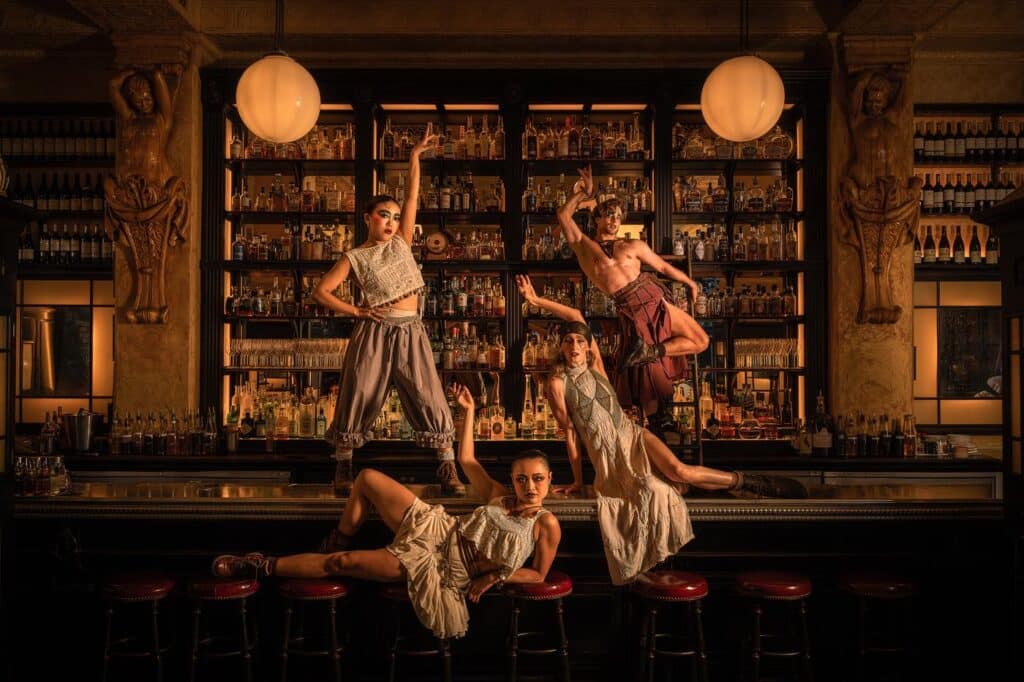 Four Cabaret performers posed across the bar at Balthazar, one lying along the stools and one mid-leap in front of the backlit bottle shelves