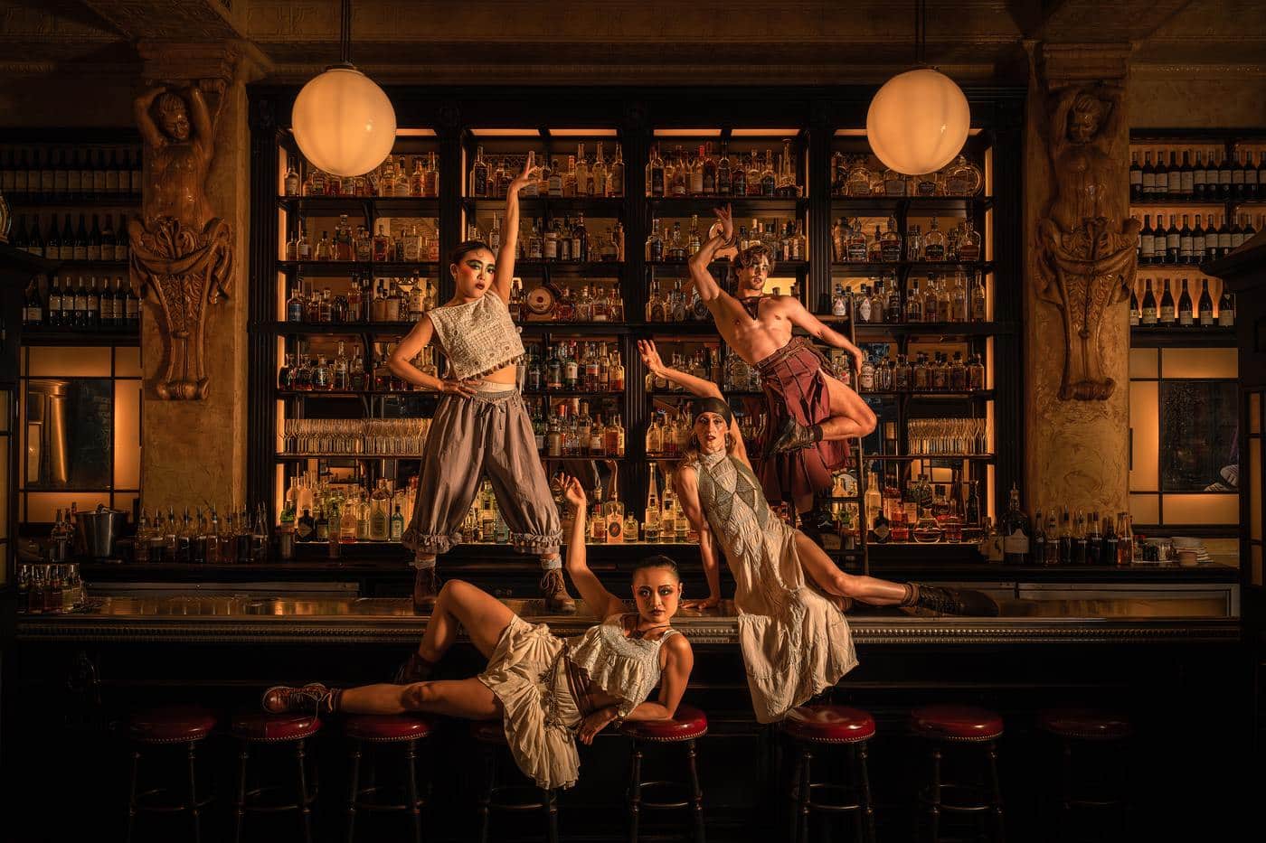 Four Cabaret performers posed across the bar at Balthazar, one lying along the stools and one mid-leap in front of the backlit bottle shelves