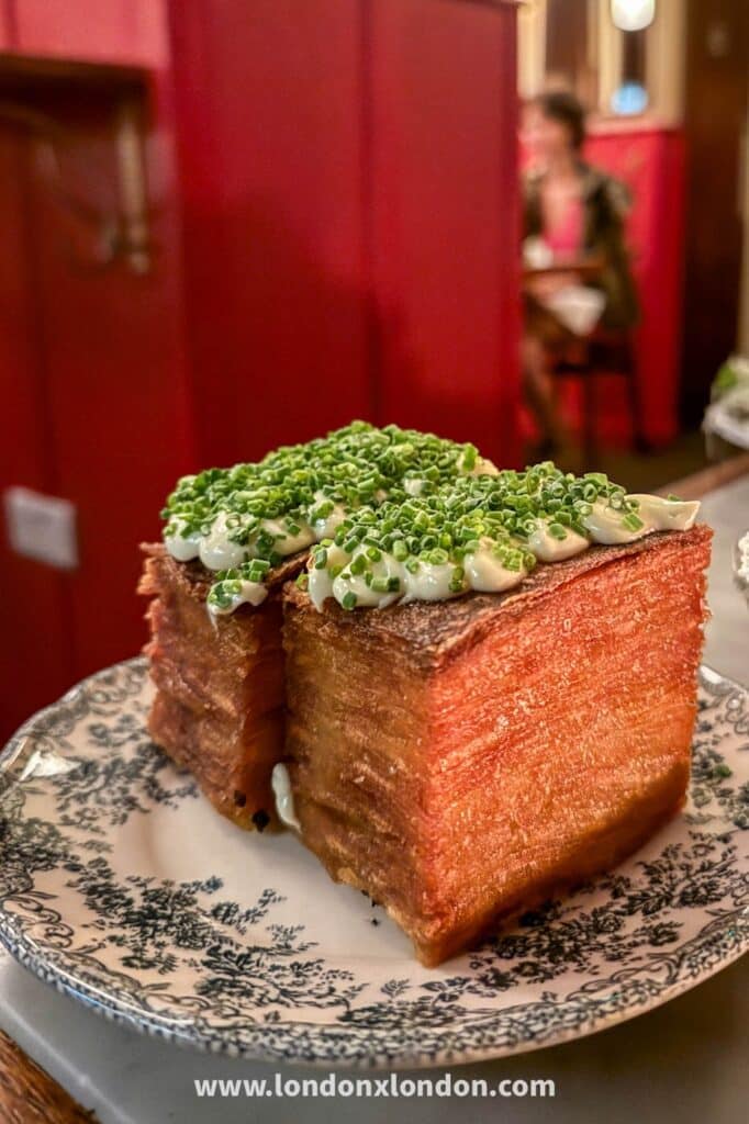 A deep-fried brick of many-layered potato topped with crème fraîche and a thick layer of chopped chives, on a green and white transferware plate at Camille