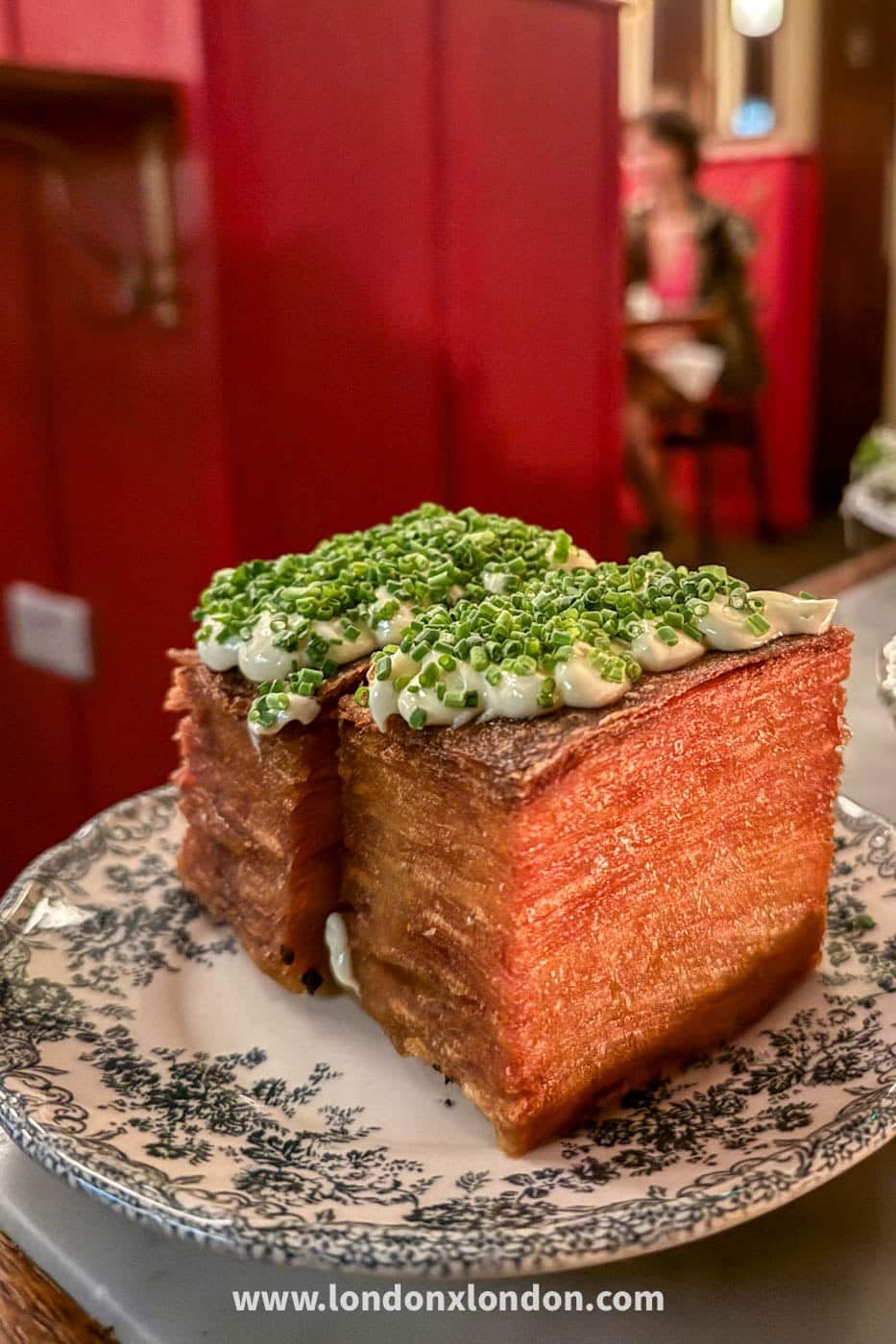 A deep-fried brick of many-layered potato topped with creme fraiche and a thick layer of chopped chives, on a green and white transferware plate at Camille