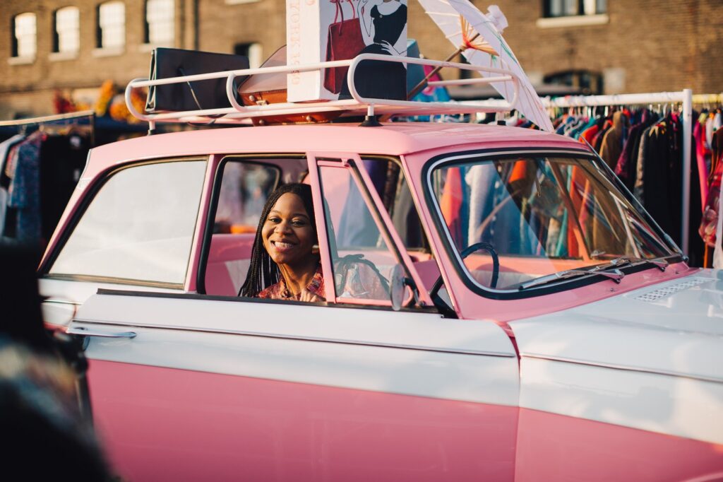 A woman smiling from the driver's seat of a pink and white classic car surrounded by clothes rails