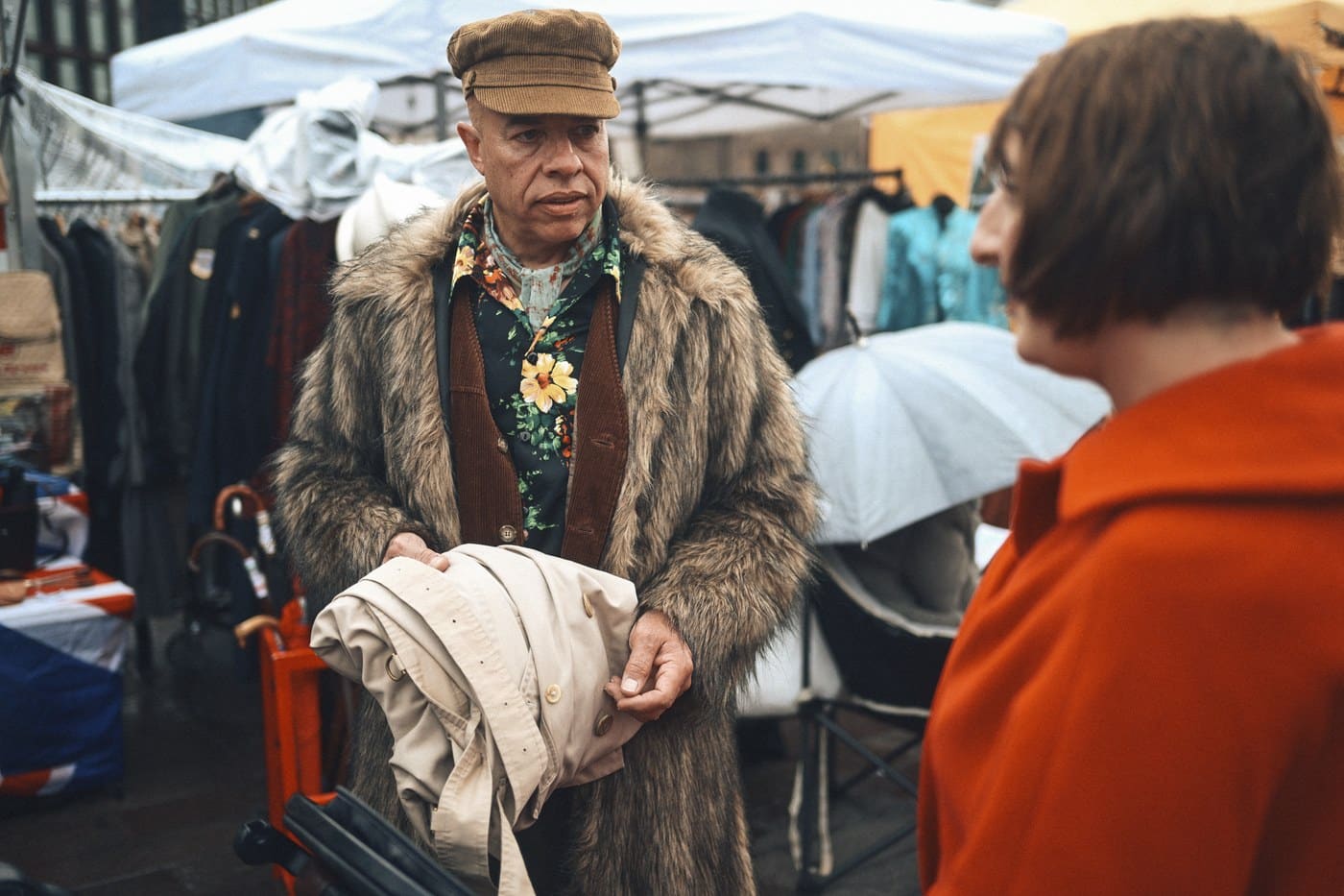 A man in a fur coat and corduroy cap holding a folded trench coat while talking to a customer
