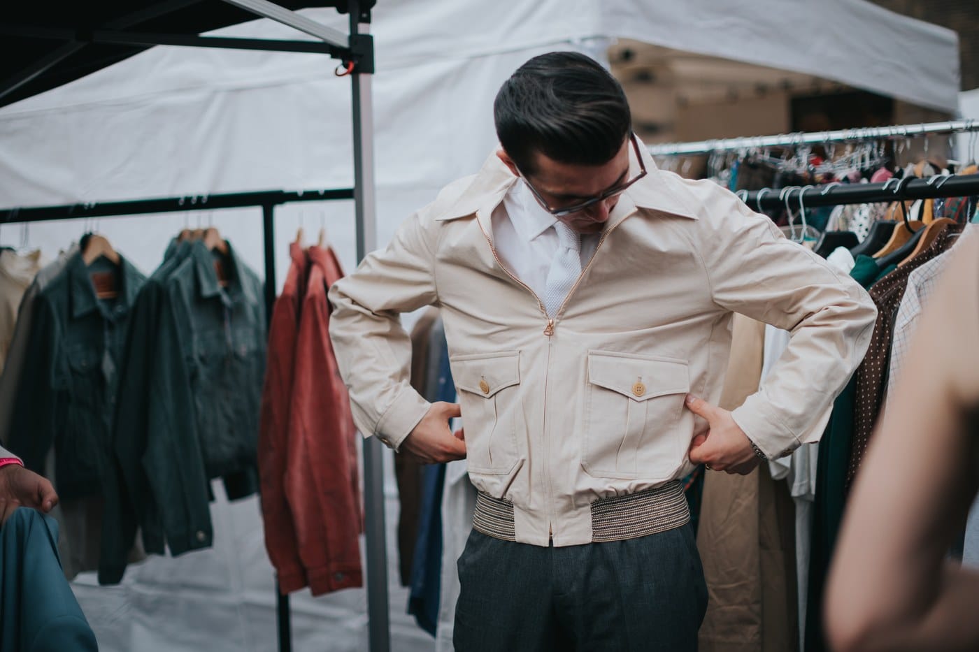 A man in glasses and a tie trying on a cream vintage bomber jacket at a market stall