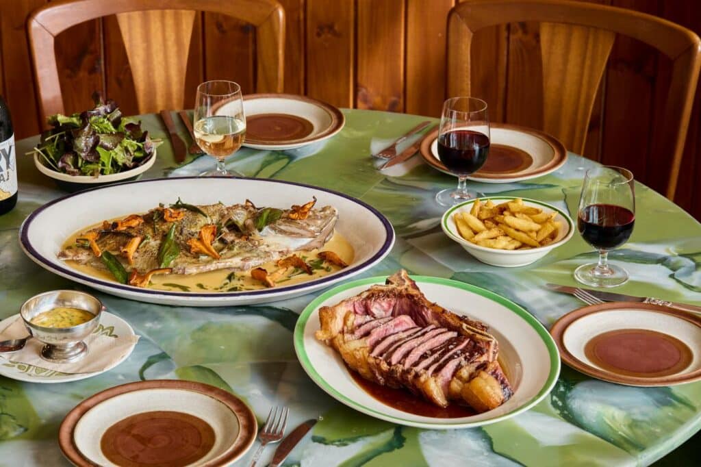 A green marble table laid with a whole roast fish in sauce with chanterelles, a sliced bone-in steak, chips, salad, bearnaise and glasses of wine