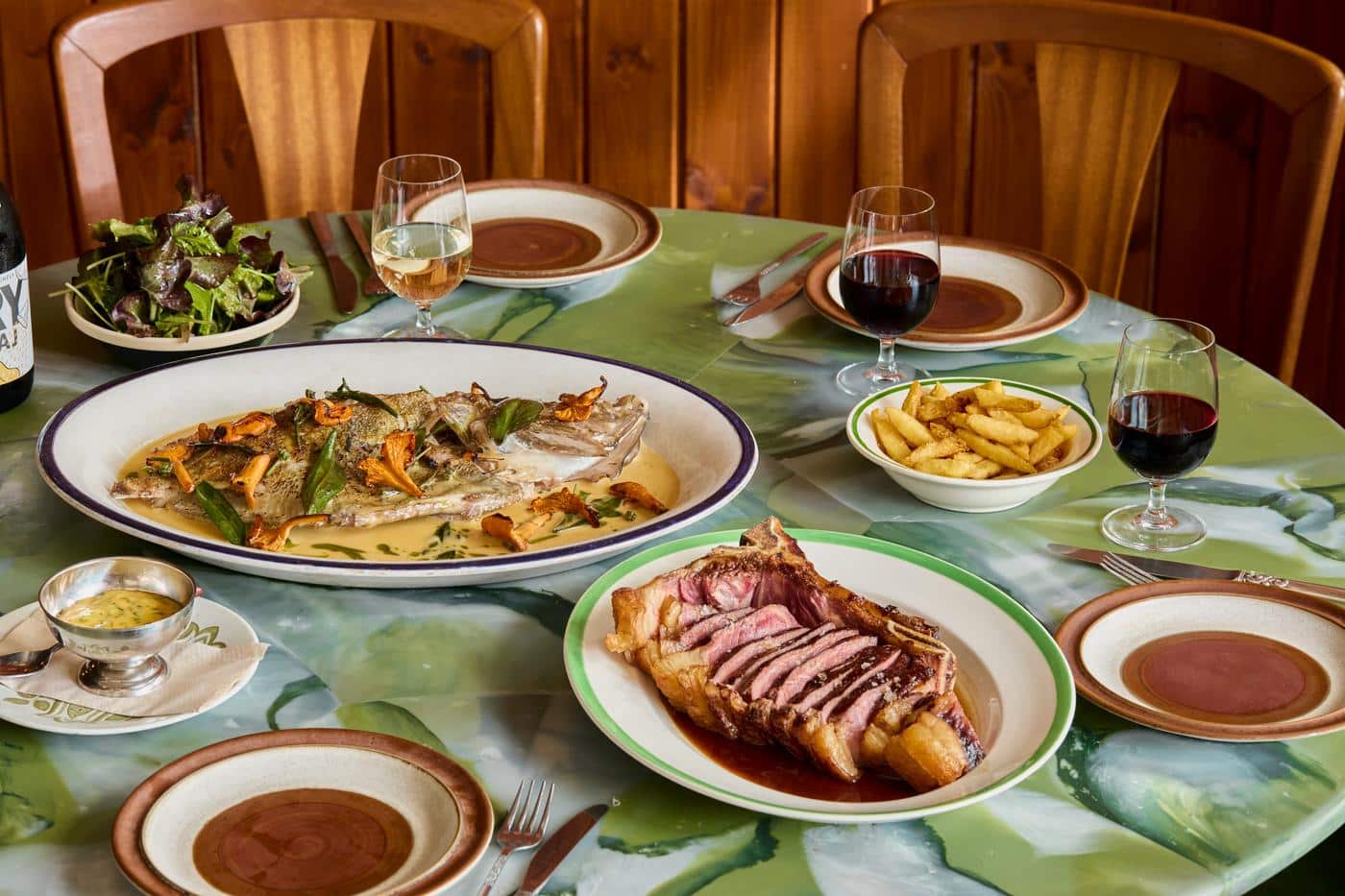 A green marble table laid with a whole roast fish in sauce with chanterelles, a sliced bone-in steak, chips, salad, bearnaise and glasses of wine