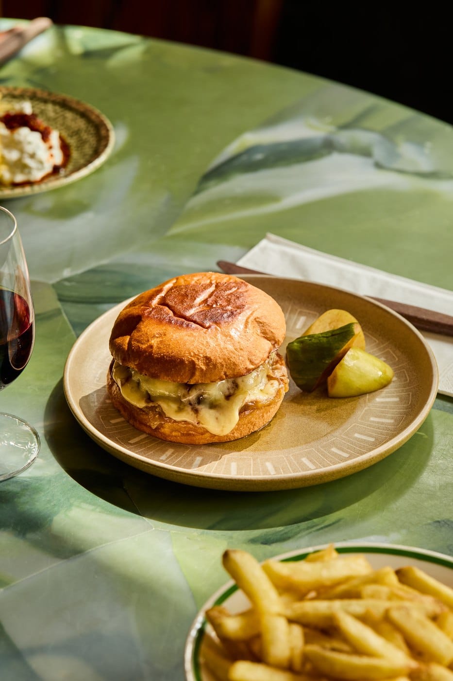 A toasted cheese sandwich oozing melted cheese on a stoneware plate with pickles, beside a glass of red wine and a bowl of chips
