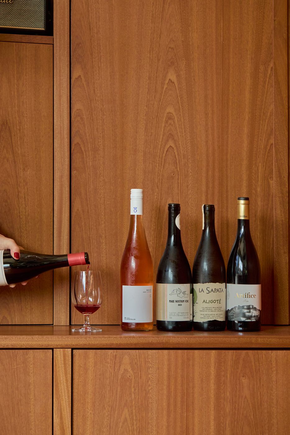 A row of low-intervention wine bottles on a wooden cabinet shelf, with red wine being poured into a glass