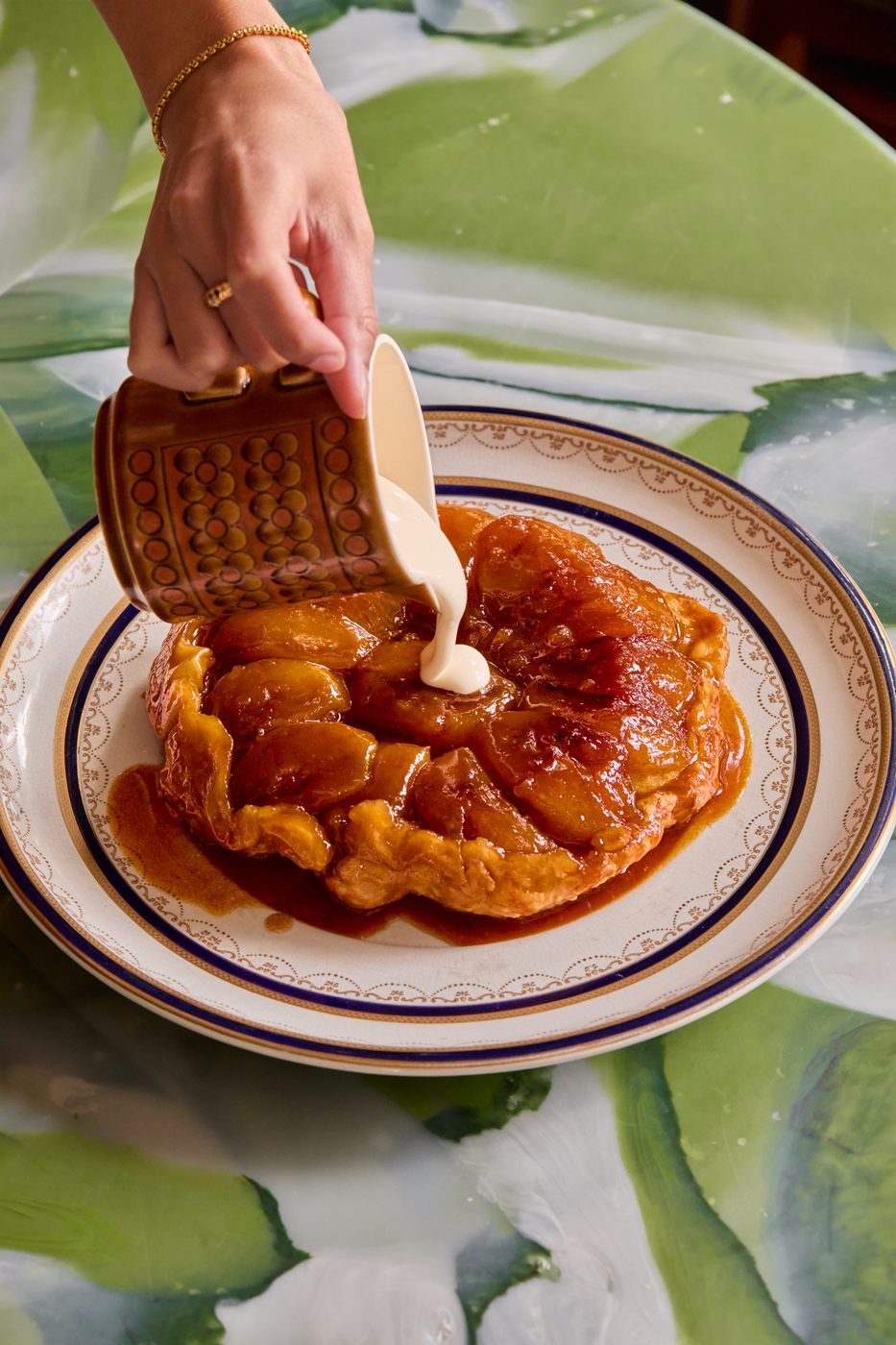 Pouring cream from a retro brown jug over an apple tarte tatin on a gold-rimmed plate, on a green marble table